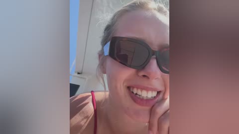 Video of a smiling blonde woman with fair skin, wearing red spaghetti straps and dark sunglasses, leaning against a white structure on a sunny day.