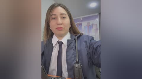 Video of a young Caucasian woman with light skin and straight brown hair, wearing a white dress shirt, dark tie, and navy blue blazer, holding a badge, in a blurred, dimly lit office setting.