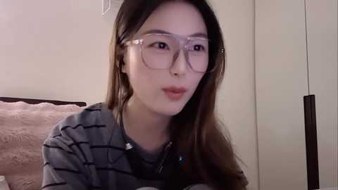 Video of an Asian woman with long brown hair, fair skin, and round glasses, wearing a striped gray shirt, leaning against a pink, fluffy pillow in a beige room.
