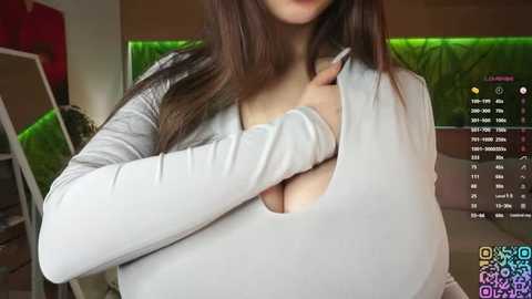 Video of a woman with long brown hair, wearing a light grey ribbed top, revealing cleavage. Background shows green aquarium and a wooden staircase.