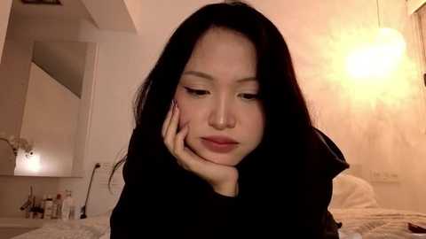 Video of an Asian woman with long black hair, fair skin, and a contemplative expression, resting her head on her hand. Background shows a softly lit room with beige walls, a lamp, and a bed.