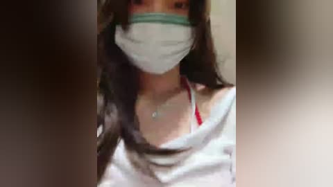 Video of a woman with long dark hair, wearing a white T-shirt, red bra, and a surgical mask, indoors against a blurred background.