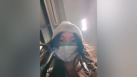Video of a woman with long dark hair, wearing a white face mask and a white hat, standing indoors with a blurry background, capturing a candid moment.