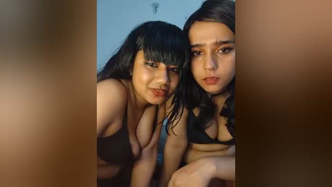 Video of two young women with medium skin tones, dark hair, and dark makeup, wearing black lingerie, leaning forward, with a blurred, nude male figure in the background.