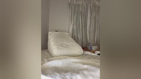Video of a serene bedroom with a white, textured, rectangular pillow on a plush, white, circular rug. Behind, a window with sheer white curtains diffuses natural light. A small table with various items is visible in the background.