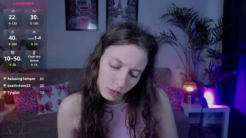 Video of a young woman with long, wavy brown hair, wearing a pink top, sitting on a couch in a dimly lit room with purple and blue lighting.