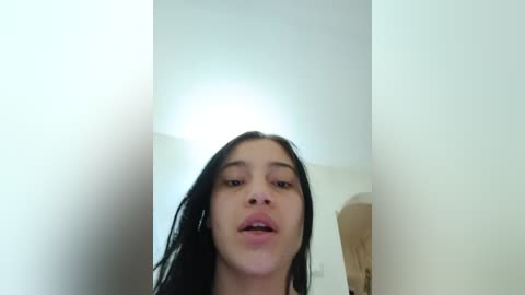 Video of a young woman with long black hair and light skin, partially obscured by a white wall, looking slightly to the right with a neutral expression.