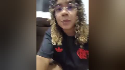 Video of a woman with curly, shoulder-length hair and glasses, wearing a black Adidas shirt with red accents. She sits on a brown leather couch in a dimly lit room.