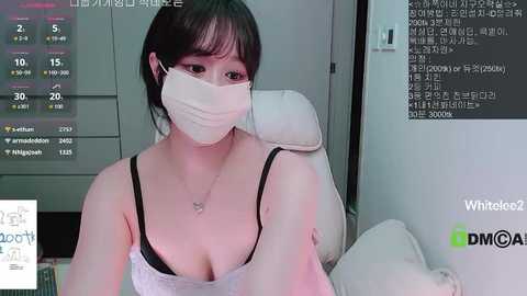 Video of a young Asian woman with short black hair, wearing a white mask, black lace bra, and a white top, sitting in a modern white chair in a dimly-lit room with a whiteboard and a computer screen displaying a calendar.