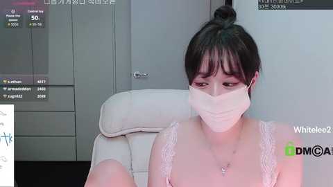 Video of a young Asian woman with short black hair in a white lace bra, wearing a face mask, sitting on a beige chair in a dimly lit room.