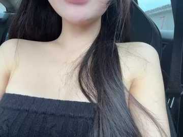 A video of a young Asian woman with long, straight black hair, partially covering her face. She is wearing a black strapless dress. The background shows the interior of a car with a grey seat.