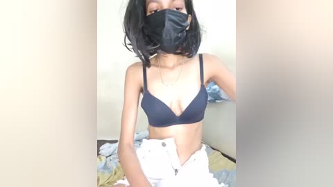 Media: Video of a young woman with short black hair, wearing a black bra, face mask, and white pants, sitting on a bed with a beige blanket.
