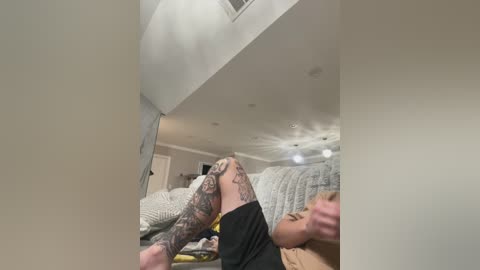 Video of a tattooed man with muscular legs in a black shirt, lying on a beige bed with a grey quilt, in a dimly lit bedroom with white walls and a ceiling fan.