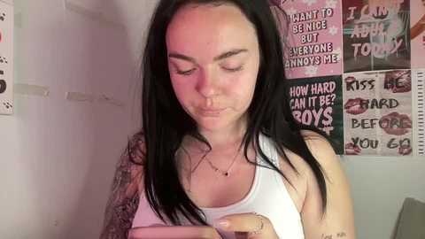 A video of a young woman with long black hair, wearing a white tank top, standing in a room with pink and black graffiti walls.