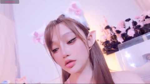 Video of a young Asian woman with fair skin, wearing cat ear headbands and pink makeup, smiling slightly, in a softly lit room with white walls and a floral arrangement.