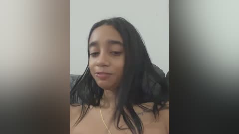 Video of a young woman with medium-brown skin and long, straight black hair, wearing a gold necklace, in a neutral expression. Background is blurred, focusing on her face.