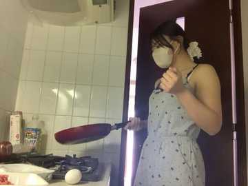 A video of an Asian woman in a light blue patterned dress, cooking with a spatula in a small, tiled kitchen. She has a white flower in her hair and wears a mask.