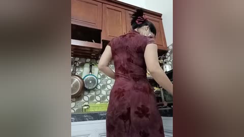 Video of a woman in a maroon velvet dress, facing away, in a kitchen with wooden cabinets, metallic hanging pots, and a tiled backsplash.