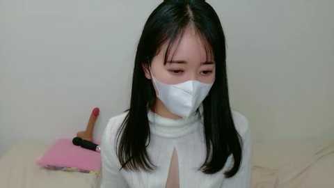Video of an East Asian woman with long black hair, wearing a white face mask and white turtleneck, holding a pink broomstick, in a minimalistic, light-colored room.