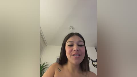 Video of a young, light-skinned Asian woman with straight black hair, smiling, topless, standing in a narrow, white-walled room with a ceiling light and a potted plant in the background.