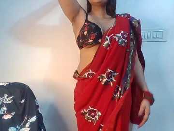 Video of a woman with medium skin tone, wearing a red sari with floral embroidery and a black floral bra, standing in a room with a floral-patterned black chair and a light gray wall.