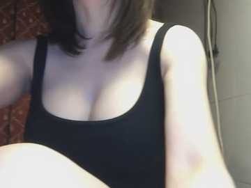 Video of a woman with fair skin and dark shoulder-length hair, wearing a black tank top that reveals cleavage, seated in a bathroom with tiled walls and a red-patterned towel.