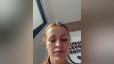 Video of a woman with fair skin and light brown hair, wearing a brown jacket, standing indoors near a window with \"Coffee\" text on a poster.