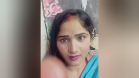 A video of a young woman with medium brown skin, wearing a blue sari, with a red dot on her forehead, against a blurred background featuring a pink flower.