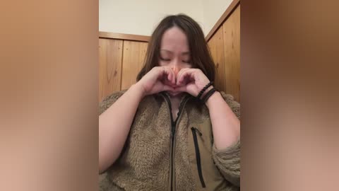 Video of a young Asian woman with straight, medium-length brown hair, wearing a brown, fuzzy jacket, making a heart shape with her hands over her face, indoors against wooden walls.