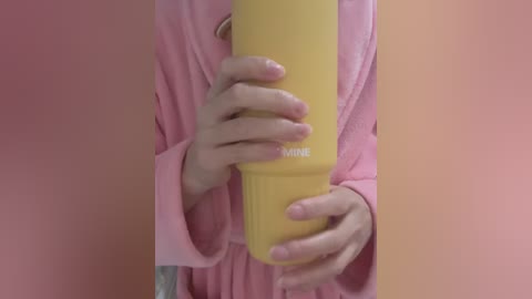 A video of a person holding a yellow water bottle with their hands. They are wearing a light pink robe with long sleeves. The background is blurred and features a soft pink hue.