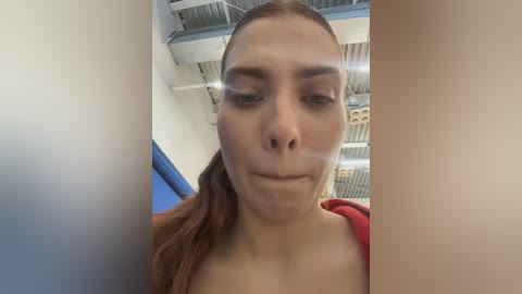 Video of a fair-skinned woman with brown hair pulled back, wearing a red top, in an industrial setting with metal beams and pipes.