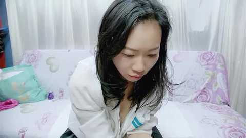 Video of an Asian woman with long black hair, wearing a white jacket, sitting on a bed with pastel floral sheets, looking down thoughtfully.