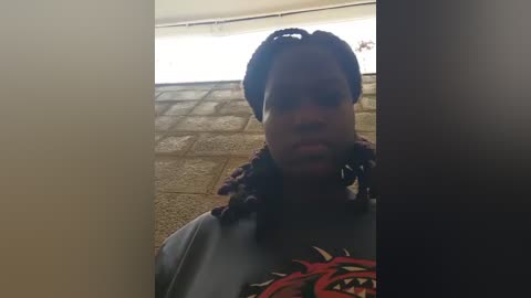 Video of a young Black woman with braided hair, wearing a graphic t-shirt, standing indoors near a tiled wall with a white ceiling.