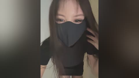 Video of an East Asian woman with long black hair, wearing a black mask and crop top, standing in a dimly lit room.