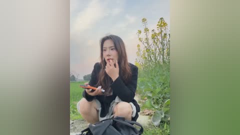 Video of an Asian woman with long, dark hair, wearing a black top and shorts, squatting in a field, holding a phone, biting her fingers, surrounded by tall sunflowers.