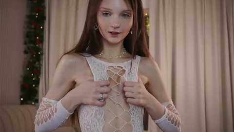 Video of a fair-skinned, slender woman with long brown hair, wearing a white lace dress with crisscross straps, standing indoors with beige curtains and a decorated Christmas tree in the background.