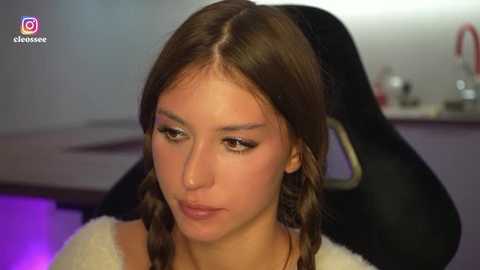 Media: Video of a young, fair-skinned woman with brown hair in braids, wearing makeup, seated on a black gaming chair in a dimly lit room.