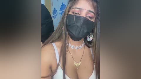 Video of a young woman with light brown skin, long brown hair, wearing a black face mask, white spaghetti strap top, and a silver necklace with a butterfly pendant, seated in a room with blue and white patterned wall.