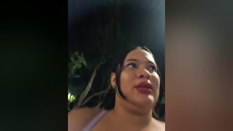 A video of a young woman with dark skin, dark hair, and a plump physique, wearing a white tank top, looking upward, surrounded by dark foliage.