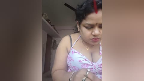 A video of an Indian woman with medium skin tone, short black hair, wearing a floral-patterned bra, a gold chain, and a red bindi. She is in a dimly lit room with a shelf and a door in the background.