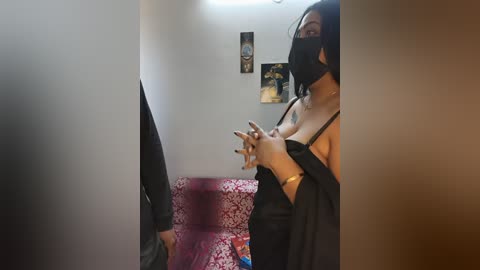 Video of a woman in a black mask and camisole, standing in a dimly-lit room with a red chair, wall posters, and a blurred figure.