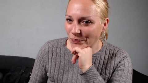 Video of a middle-aged, fair-skinned woman with blonde hair tied back, wearing a gray knitted sweater, resting her chin on her hand, in a neutral indoor setting.