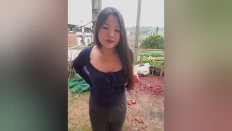 Video of a young Asian woman with long black hair, wearing a dark blue top and olive green pants, standing in a rural setting with greenery, red dirt, and scattered objects.