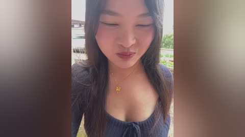 Video of an Asian woman with long black hair, wearing a low-cut navy blue top, standing outdoors in daylight. She has a serene expression, with a subtle smile.