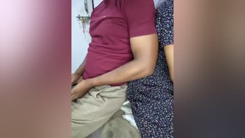 Video of a person in a maroon shirt and beige pants, embracing a woman in a blue floral dress. Background shows a white wall with indistinct objects.