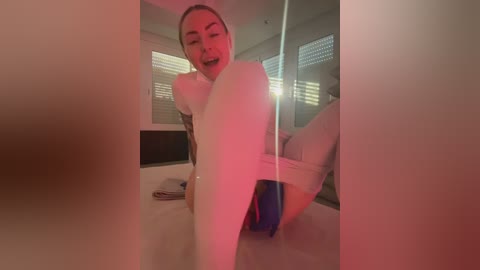 A video of a blonde woman with a medium build, wearing a white bodysuit and a blue thong, sitting on a white bed with blinds in the background, under red lighting.