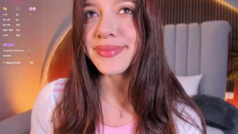 Video of a smiling young woman with long, brown hair, light skin, wearing a white top, standing in a modern bedroom with a gray upholstered headboard.