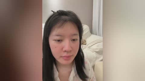 Video of an East Asian woman with long black hair and light skin, wearing a floral-patterned robe, sitting in a beige room with a cream couch and a white wall.
