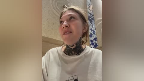 Video of a young woman with blonde hair, wearing a white t-shirt, and a detailed black tattoo around her neck, standing in a bathroom with a floral shower curtain and beige ceiling.