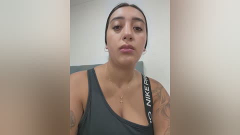 Video of a young woman with medium skin tone, dark hair, wearing a black Nike tank top, and visible tattoos. She has a neutral expression and is indoors with a blurred background.
